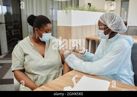 Ernsthafte afroamerikanische Frau in medizinischer Maske, die einen Arzt ansieht, der ihre Dosis Impfstoff gegen Covid-19 verabreichte Stockfoto