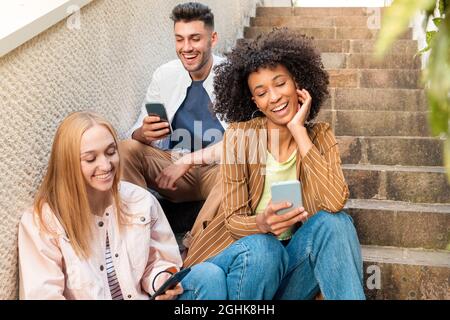 Fröhlicher, multirassischer junger Mann und Frauen in legerer Kleidung, lächelnd und mit Freude auf Handys surfen, während sie auf der Straße auf der Treppe sitzen Stockfoto