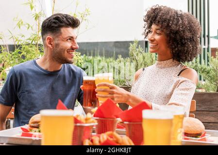 Fröhliches junges, multirassisches Paar, das in einem Restaurant im Freien ein Sommerwochenende gemeinsam genießt und den Urlaub feiert, klirrrende Gläser Bier Stockfoto