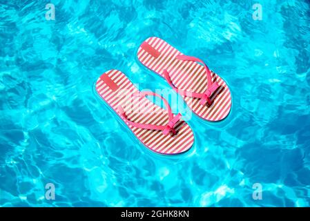 Ein Paar rot-weiß gestreifte Flip Flops, die im Sommer in einem funkelnden, sonnendurchfluteten Swimmingpool auf dem blauen Wasser schwimmen Stockfoto