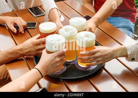 Eine Gruppe anonymer Personen, die volle Gläser schaumiges Bier vom Tablett nehmen, während sie sich während des Treffs mit Mobiltelefonen am Holztisch treffen Stockfoto