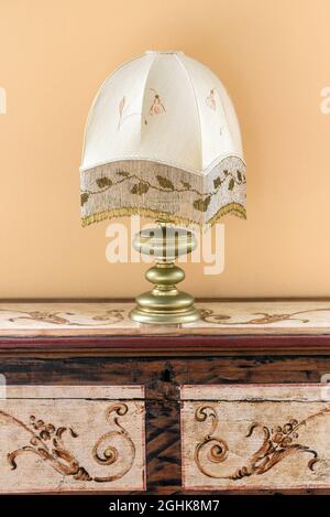 Altmodische Tischleuchte mit gemustertem Lampenschirm auf einem Vintage-Holzschrank mit verzierten Elementen an der beigefarbenen Wand im Zimmer Stockfoto