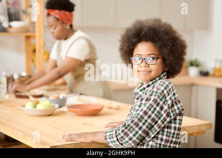 Netter kleiner Junge, der am hölzernen Küchentisch gegen seine Mutter steht, die hausgemachtes Gebäck kocht Stockfoto