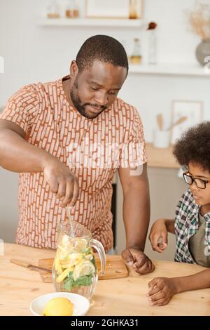 Der junge Vater mischte Zitronenscheiben und Schnitt Minzblätter in einen Krug, während er seinem kleinen Sohn zeigte, wie man Limonade macht Stockfoto