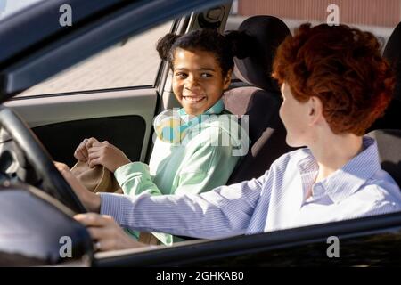 Glückliches Mixed-Race-Mädchen, das junge Frau ansieht, während beide mit dem Auto zur Schule gehen Stockfoto