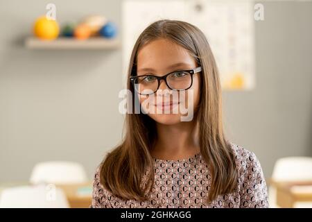 Nette Schülerin der Sekundarstufe in einer Brille, die im Klassenzimmer auf die Kamera schaut Stockfoto