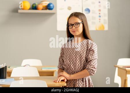 Attraktives Schulmädchen in Kleid und Brille, das an ihrem Schreibtisch gegen Bilder und Modelle von Planeten steht Stockfoto
