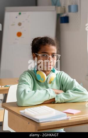 Nettes ernsthaftes Schulmädchen in Brillen, das an ihrem Schreibtisch sitzt und beim Unterricht die Kamera anschaut Stockfoto