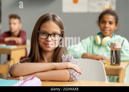 Nettes Schulmädchen in Brillen, das Sie anschaut, während Sie am Schreibtisch vor der Kamera sitzen Stockfoto