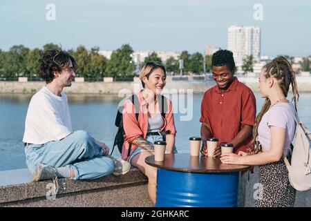 Eine Gruppe von glücklichen jungen interkulturellen Freunden, die am Flussufer etwas trinken und miteinander interagieren Stockfoto