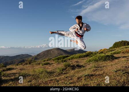 Karate Boy in weißem Kimono tritt in die Luft, während er im Freien Kampfkunst übt. Stockfoto