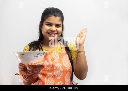 Eine hübsche indische Hausfrau, die Kochschürze und Ofenhandschuhe mit einer Schüssel in der Hand trägt, lächelt auf weißem Hintergrund Stockfoto