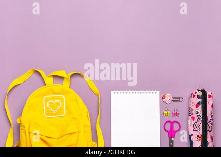 Zurück zur Schule, Bildungskonzept. Gelber Rucksack mit Schulbedarf - Notizbuch, Stifte, Clips, Schere isoliert auf violettem Hintergrund Draufsicht Copy Stockfoto