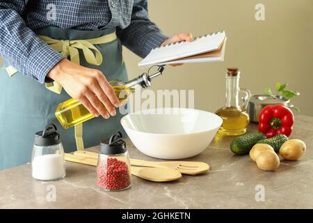 Eine Frau in einer Schürze hält ein Rezeptbuch und eine Flasche Öl und kocht Stockfoto