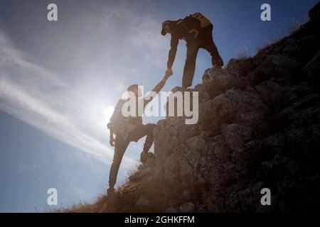 Hilfe- und Hilfekonzept. Silhouetten von zwei Menschen, die Berge besteigen und gegen den blauen Himmel helfen. Gehen Sie in den Wolken. Discovery Travel Direc Stockfoto