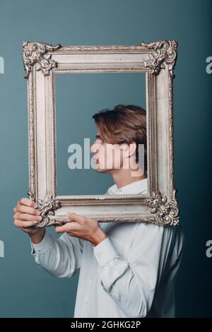Millennial junger Mann mit blonden Haaren auf vergoldeten Bilderrahmen-Profilporträt. Isoliert auf blauem Hintergrund im Studio. Stockfoto