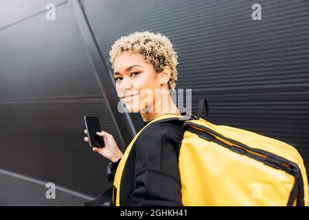 Porträt einer lächelnden Zustellerin mit Tasche, die auf einem Bürgersteig steht und das Smartphone hält Stockfoto