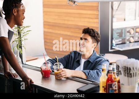 Junger Mann, der Saft auf einer Theke eines Food-Trucks hält und die Verkäuferin ansieht Stockfoto