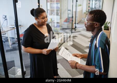 Positive Geschäftsfrau mit einem Bericht in der Hand, der ihrem Kollegen, den sie im Büroflur getroffen hat, Fragen stellte Stockfoto