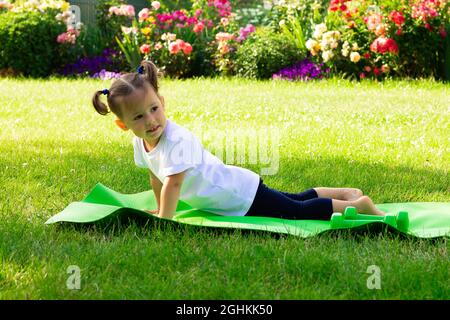 Kleines süßes Mädchen 1-3 in einem weißen T-Shirt geht in für Sport auf einer grünen Matte auf dem Gras, vor einem Hintergrund von Blumen Stockfoto