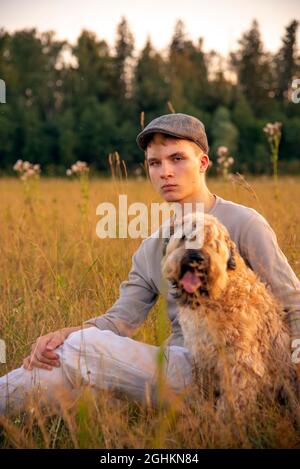 Ein 18-jähriger Junge mit Mütze sitzt an einem Sommerabend mit einem Hund auf einem Feld im dicken Gras. Stockfoto