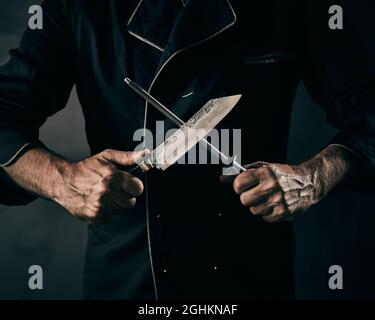 Der Koch schärft ein Messer oder einen Spaltmesser auf einem altmodischen Handheld in einem schattigen Licht in Nahaufnahme eine Metallfeile an seinen Händen Stockfoto