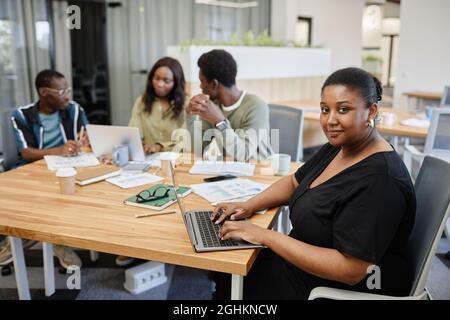 Positive schwarze Unternehmerin, die am Laptop arbeitet, als ihre Kollegen eine Briefing-Sitzung abhalten Stockfoto