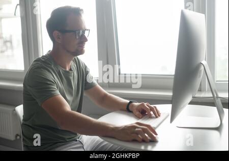 Schöner junger Hipster-Freiberufler mit Bart in Brille, der am Tisch sitzt und auf den PC-Monitor schaut, lächelnder Kerl, der gerne von zu Hause aus arbeitet, Nachrichten tippt und E-Mails beantwortet Stockfoto