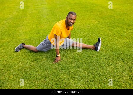 Lächelnder afroamerikanischer Mann in stilvoller Sportkleidung, der auf dem Gras sitzt und versucht, die Beine gespalten und zu strecken. Der athletische Mann arbeitet, macht Stockfoto