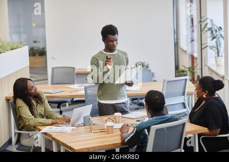 Junger Geschäftsmann, der die Ergebnisse seiner Marktforschung bei einem Treffen mit Kollegen vorliest Stockfoto