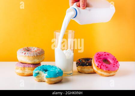 Bunte Donuts mit süßer Marmelade und Erdnussbutterfüllung auf grauem Tisch. Mann Hand gießt Pflanzenmilch auf das Glas. Vorderansicht gelb Stockfoto