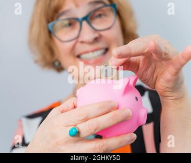 Eine ältere Frau mit Brille legt eine Münze in ein Sparschwein auf weißem Hintergrund Stockfoto