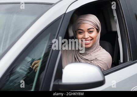 Fröhliche islamisch-afroamerikanische Fahrerin in Hijab sitzt im neuen Auto am Lenkrad und schaut auf die Kamera Stockfoto