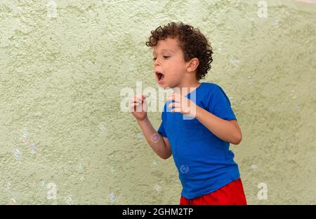 Lockiger Junge mit Sommersprossen, die im Hinterhof mit Seifenblasen spielen. Stockfoto