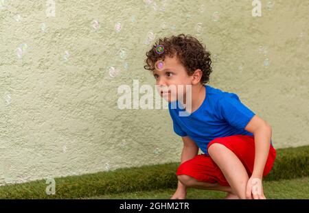 Lockiger Junge mit Sommersprossen, die im Hinterhof mit Seifenblasen spielen. Stockfoto