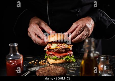 Koch bereitet einen Gourmet Big Boy Double Burger mit kompletten Hauszutaten wie Käse, Speck, Salatzutaten und Senf zu, um das geröstete Brötchen zu servieren Stockfoto