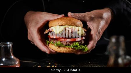 Mann hält einen köstlichen Double Beef Burger mit Senfsauce, Speck, Salatzutaten und würziger Hacksteaks auf einem gerösteten Brötchen für die Speisekarte Adverti Stockfoto