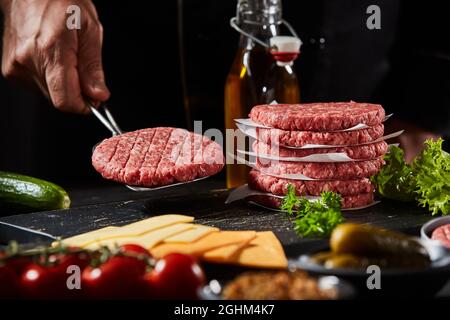 Der Koch hebt ein frisch zubereitetes hausgemachtes Rinderpastete auf einem Spatel, während er Cheeseburger in einem Restaurant zubereitet, das im niedrigen Winkel über Zutaten stand Stockfoto