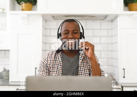 Lachender afroamerikanischer Mann, der in der Küche sitzt und mit einem Laptop und einem Headset Videoanruf macht Stockfoto