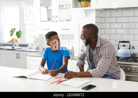 Glücklicher afroamerikanischer Vater und Sohn in der Küche, gemeinsam Hausaufgaben machen Stockfoto