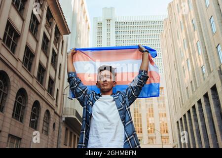 Transman, der die Transgender-Flagge im Freien aufhebt. Selbstbewusster junger Transgender-Mann, der den Stolz der Homosexuellen in der Stadt feiert. Junger Mann, der nicht dem Geschlecht entspricht, stan Stockfoto