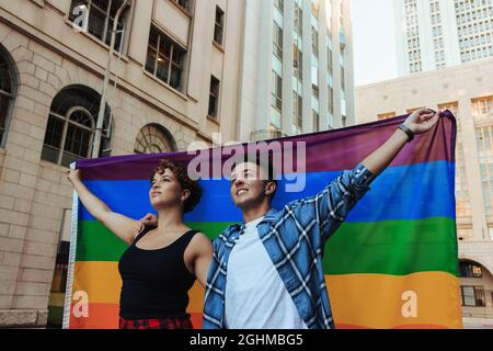 Pärchen bei einer Parade zum Stolz. Selbstbewusstes junges LGBTQ+-Paar, das die Regenbogenfahne hochhebt, während es in der Stadt zusammensteht. Junge, queere Paare feiern g Stockfoto