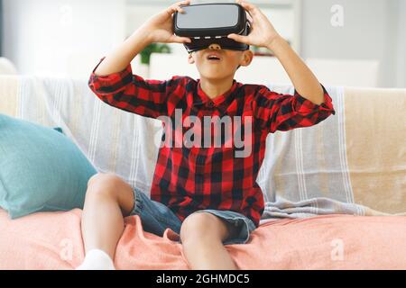 Lächelnder asiatischer Junge, der auf der Couch sitzt und ein vr-Headset benutzt Stockfoto