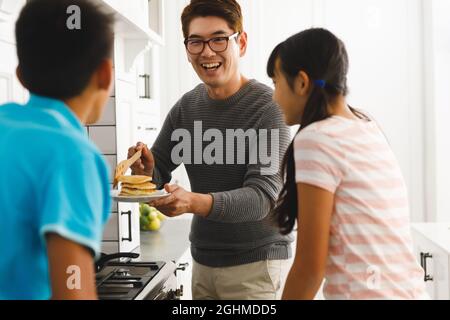 Lächelnder asiatischer Vater hält Pfannkuchen und bereitet das Frühstück mit Sohn und Tochter in der Küche vor Stockfoto