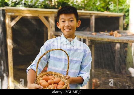 Porträt eines asiatischen Jungen, der lächelt und einen Korb hält und Eier aus dem Hühnerstall im Garten sammelt Stockfoto