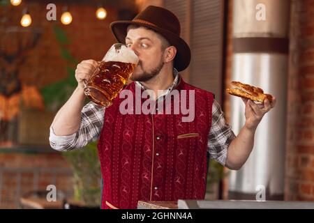 Porträt eines glücklichen bärtigen Mannes in traditioneller bayerischer Festtracht, Hut und roter Weste, der während des Bierfestes in Bar, Café, Pub Bier und Snack verkostet Stockfoto