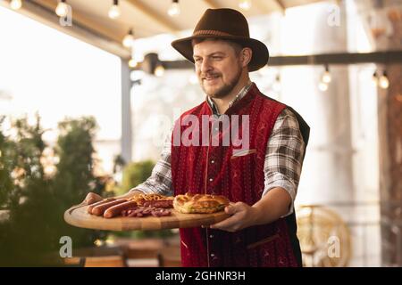 Fröhlicher lächelnder Mann, Kellner in traditioneller bayerischer Tracht, der ein rundes Holztablett mit festlichem Essen, Brezel und Würstchen hält. Oktoberfest Stockfoto