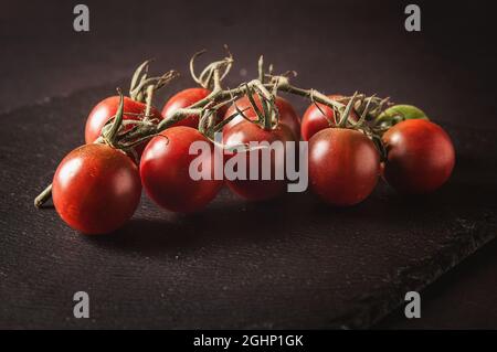 Ein paar frische Kirschtomaten auf einem Zweig befinden sich auf einem schwarzen Servierbrett auf dunklem Hintergrund Stockfoto