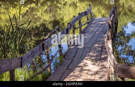 Eine Holzplankenbrücke auf einem See mit Reflexionen auf der Wasseroberfläche. Seeufer mit Schilf und üppigem Gras bedeckt. Meditation, Entspannung in der Natur, Stockfoto