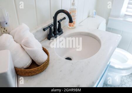 Ein Korb mit organisierten, sauberen, rollenden weißen Handtüchern auf einer Badezimmertheke in einem Gästebad neben einem Waschbecken und einer Toilette. Stockfoto